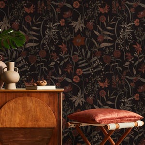 May include: A dark floral wallpaper with red and tan flowers and foliage. A wooden cabinet with a round door and two vases sits in front of the wallpaper. A small, wooden folding stool with a red cushion is in the foreground.