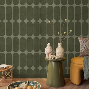 Pode incluir: Um interior com uma parede verde escuro com um padrão oval. Uma pequena mesa verde suporta um vaso com flores secas e uma escultura. Uma tigela tecida com cabaças está no chão e uma pequena banqueta de vime é visível.