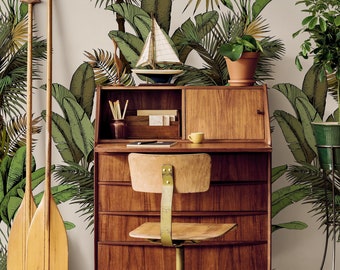 Può includere: Una scrivania in legno con una carta da parati a stampa tropicale con foglie di palma e foglie di banana. La scrivania ha un cassetto superiore, tre cassetti sotto e una sedia con seduta e schienale in legno. La scrivania si trova in una stanza con una parete bianca e una pianta verde in un vaso.