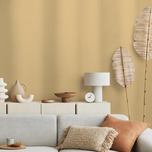 Può includere: Interno di un soggiorno con un divano beige chiaro, cuscini decorativi e un tavolino in legno. Un mobile bianco espone vari vasi in ceramica, una lampada e un piccolo orologio. Due grandi decorazioni di foglie secche sullo sfondo.