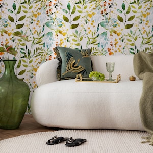 May include: A white couch with a green throw blanket and a green vase. The couch has a floral patterned wallpaper behind it. There is a tray with a glass of water and a bowl of grapes on the couch.