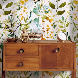 May include: A wooden dresser with two drawers and a cabinet door. The dresser has gold-colored knobs. The dresser is in front of a white wall with a watercolor floral pattern in shades of green, blue, and yellow.