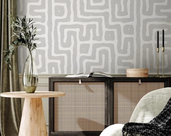 May include: A room interior featuring a light gray wall with a white, abstract maze pattern. A wooden cabinet with woven panels and a round wooden side table are in the foreground. A vase with greenery and a gold candle holder are also visible.