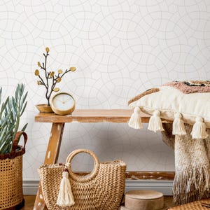 May include: A wooden bench displays a woven straw tote bag with a tassel, a decorative clock, and a small plant. A woven basket with a handle and a decorative pillow with tassels are also visible. The background features a white wall with a geometric pattern.