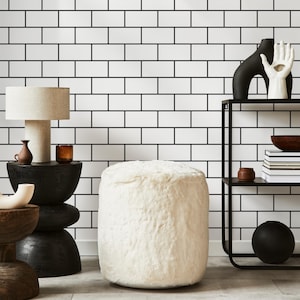 May include: A contemporary interior scene with a white faux fur ottoman, a black side table with a lamp, and a black metal shelf with decorative items. The backdrop is a white tiled wall with black grout.