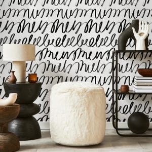 May include: A room interior featuring a white faux fur ottoman, a black side table with a lamp, and a black metal shelf. The background is a white wall with black cursive writing. Decorative items include vases and a black sphere.
