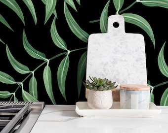 Op de afbeelding: Een witte marmeren snijplank, een kleine vetplant in een witte pot en een decoratieve pot met een houten deksel staan op een wit dienblad. De achtergrond heeft groene bladerpatronen op een zwarte achtergrond.