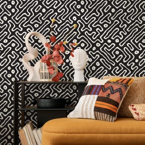 May include: A decorative interior scene featuring a black and white patterned wallpaper backdrop. A black shelf displays a white sculpture, a vase with red leaves, and a white bust. A patchwork pillow rests on a yellow sofa, with a woven pillow nearby.
