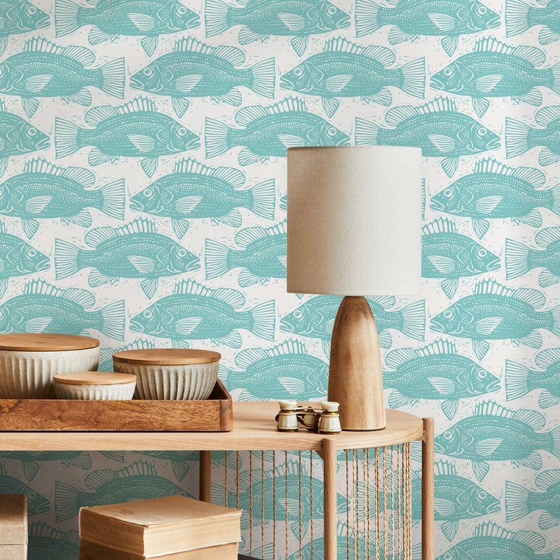 May include: A teal blue and white wallpaper with a repeating pattern of fish. The fish are stylized and have a blocky, graphic design. The wallpaper is shown in a home setting with a lamp and a wooden table.