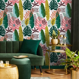 May include: A green velvet sofa with a green velvet ottoman in front of it. The sofa and ottoman are in a living room with a white wall covered in a tropical leaf wallpaper. There is a gold metal side table with a plant and glasses on it.