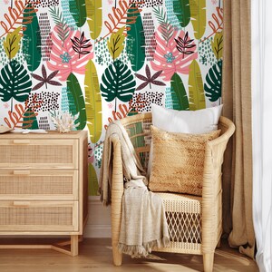May include: A wicker chair with a throw blanket and a pillow sits in front of a wall with a tropical leaf pattern wallpaper. The wallpaper features green, yellow, orange, and pink leaves.