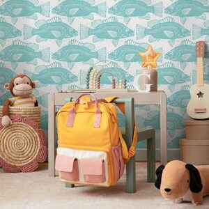 May include: A yellow backpack with pink pockets is hanging on a light green chair. A wooden table with a light green chair is in front of a white wall with a blue fish pattern. A stuffed monkey is sitting on a woven basket. A brown stuffed dog is on the floor.