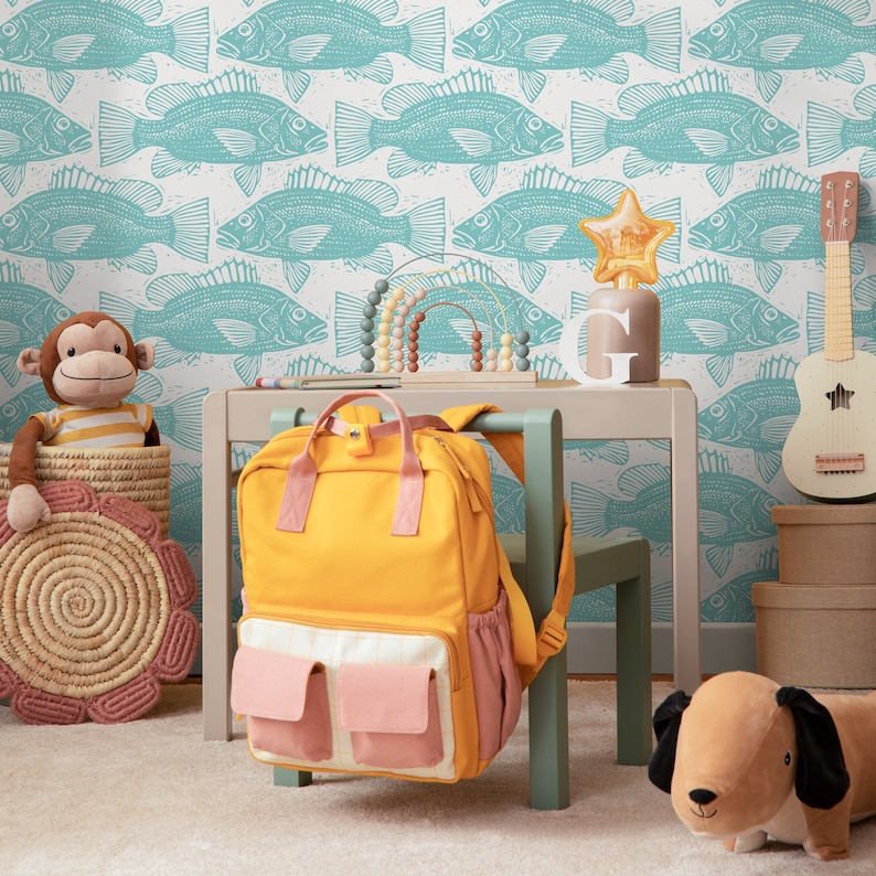 May include: A yellow backpack with pink pockets is hanging on a light green chair. A wooden table with a light green chair is in front of a white wall with a blue fish pattern. A stuffed monkey is sitting on a woven basket. A brown stuffed dog is on the floor.