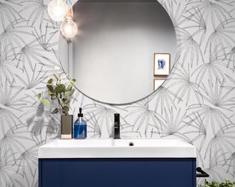 Può includere: Un bagno bianco con un mobile da bagno blu e uno specchio rotondo. Le pareti sono rivestite con una carta da parati a foglie di palma in bianco e nero. Una piccola pianta si trova sul bancone accanto a una bottiglia di sapone.