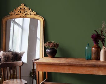 May include: A room with a dark green wall and a wooden table. A large, ornate gold mirror leans against the wall. The table holds vases with dried flowers and a blue glass bottle. A wooden chair sits near the mirror.
