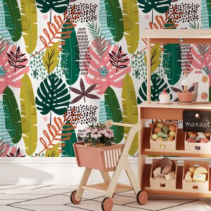 May include: A play kitchen with a chalkboard sign that says "market" and shelves stocked with toy fruits and vegetables. A pink toy shopping cart is in the foreground with a potted plant inside.