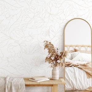 May include: A neutral-toned bedroom scene featuring a floral wallpaper design. A gold-framed arched mirror reflects a bed with white linens and a tan throw. A wooden bench holds books, a tan blanket, and a vase with dried flowers.