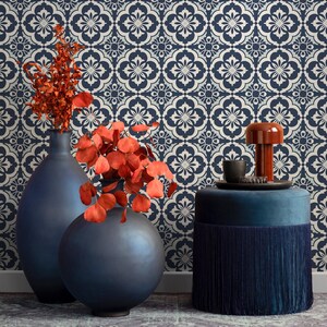 May include: A home decor scene featuring a navy blue and white patterned wallpaper backdrop. In the foreground, there are two navy blue vases with orange foliage, a blue velvet ottoman with fringe, and a small orange lamp.