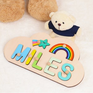 Könnte beinhalten: Ein Puzzle aus Holz in Wolkenform mit dem Namen "MILES", der in bunten Buchstaben ausgeschrieben ist. Das Puzzle ist mit einem Regenbogen, einem Stern und einer Sternschnuppe bemalt. Ein kleiner weißer Teddybär mit einem blauen Hemd sitzt neben dem Puzzle.