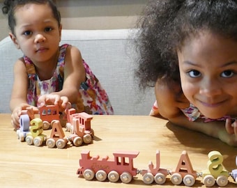 May include: Two children play with wooden train sets. One train is made up of letters spelling out the name "JASON" and the other train is made up of numbers and letters. The trains are on a light brown wooden table.