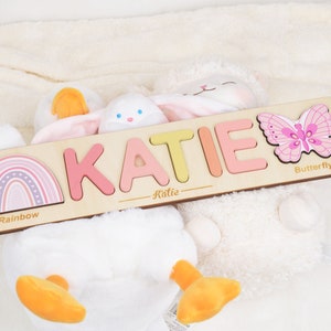 May include: A personalized wooden puzzle with the name "KATIE" spelled out in colorful letters. The puzzle also features a rainbow, a butterfly, and the words "Rainbow" and "Butterfly".