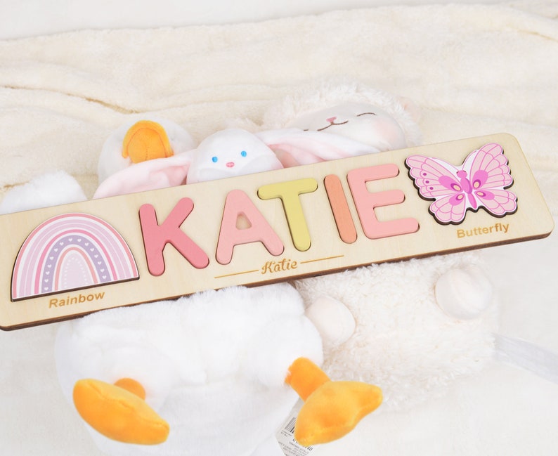 May include: A personalized wooden puzzle with the name "KATIE" spelled out in colorful letters. The puzzle also features a rainbow, a butterfly, and the words "Rainbow" and "Butterfly".