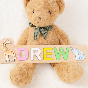 May include: A wooden name puzzle with the name "DREW" spelled out in colorful letters. The puzzle features a giraffe and an elephant on either end. A brown teddy bear is holding the puzzle.