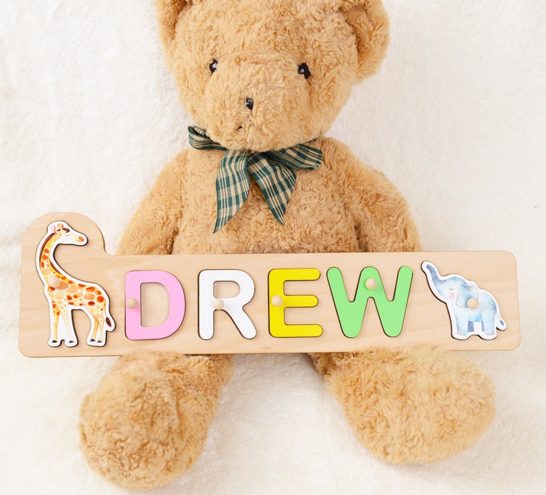 May include: A wooden name puzzle with the name "DREW" spelled out in colorful letters. The puzzle features a giraffe and an elephant on either end. A brown teddy bear is holding the puzzle.