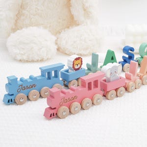 May include: Two wooden toy trains in light blue and pink, each featuring the name Jason in cursive script. The trains pull carriages carrying wooden animal figures, including a lion and an elephant, followed by individual letter blocks.