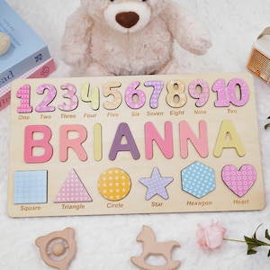 Op de afbeelding: Een rechthoekig houten puzzelbord met de cijfers &eacute;&eacute;n tot en met tien, de naam BRIANNA in grote kleurrijke letters en zes vormen, waaronder een vierkant, driehoek, cirkel, ster, zeshoek en hart, elk voorzien van de bijbehorende naam.