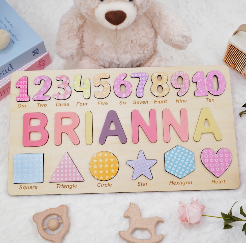 Op de afbeelding: Een rechthoekig houten puzzelbord met de cijfers &eacute;&eacute;n tot en met tien, de naam BRIANNA in grote kleurrijke letters en zes vormen, waaronder een vierkant, driehoek, cirkel, ster, zeshoek en hart, elk voorzien van de bijbehorende naam.