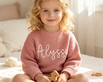 May include: A dusty rose knitted sweater featuring the name Alyssa embroidered in white cursive script across the chest. The garment has a ribbed crew neckline, long sleeves with ribbed cuffs, and a textured knit pattern throughout the fabric.
