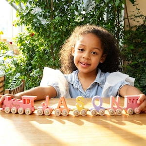 May include: A wooden toy train set featuring a pink engine with the name Jason printed on the side, followed by individual letter blocks spelling JASON in orange, yellow, purple, and pink, each mounted on small wheels with a pink caboose at the end.