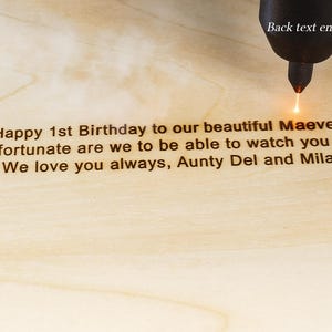 Op de afbeelding: Een lasergraveergereedschap markeert lichtgekleurd hout met de tekst: Happy 1st Birthday to our beautiful Maeve. How fortunate are we to be able to watch you grow. We love you always, Aunty Del and Mila. De tekst Back text engraving is zichtbaar.