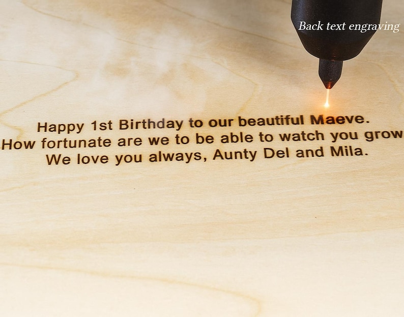 Op de afbeelding: Een lasergraveergereedschap markeert lichtgekleurd hout met de tekst: Happy 1st Birthday to our beautiful Maeve. How fortunate are we to be able to watch you grow. We love you always, Aunty Del and Mila. De tekst Back text engraving is zichtbaar.