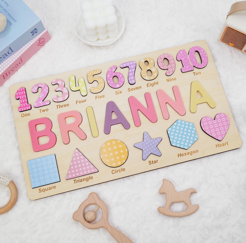Op de afbeelding: Een rechthoekig houten puzzelbord met de naam BRIANNA in grote gekleurde letters, de cijfers &eacute;&eacute;n tot en met tien met bijbehorende labels, en diverse vormen zoals een vierkant, driehoek, cirkel, ster, zeshoek en hart.