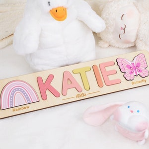 May include: Personalized wooden puzzle with the name "KATIE" spelled out in colorful letters. The puzzle also features a rainbow, a butterfly, and the words "Rainbow", "Butterfly", and "Katie".