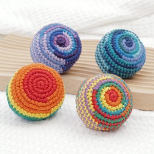 May include: Four colourful, handmade crochet balls. The balls are in shades of blue, purple, orange, green, yellow, and red. Each ball has a spiral pattern. The balls are sitting on a wooden surface.
