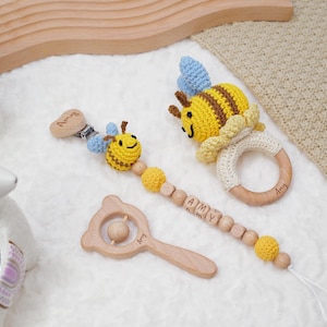 Op de afbeelding: Een houten babyratel met een berenkopontwerp, een houten bijtring met de naam "Amy" erop gegraveerd en een gehaakte bij-ratel met een geel en bruin lichaam en blauwe vleugels. De bij-ratel is bevestigd aan een houten kralenketting met gele en bruine kralen en houten letterkralen die "AMY" spellen.