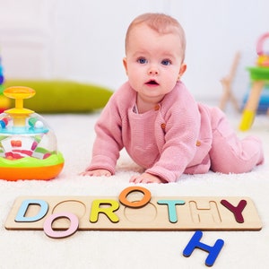 May include: A baby is playing with a wooden puzzle that spells out the name "DOROTHY". The puzzle has colorful letters and includes a few extra letters, including a blue "H".