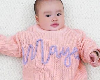 May include: A pink knitted sweater with the name "Maya" embroidered in purple on the front.