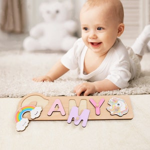 May include: A wooden puzzle with the name "AMY" spelled out in colorful letters. The puzzle includes a rainbow, cloud, and unicorn design. A baby is playing with the puzzle on a white carpet.