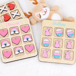 May include: Two wooden tic-tac-toe boards with personalized names, "Eloise" and "Brittany". One board has rainbow and heart shaped game pieces, the other has bunny and Easter egg shaped game pieces.