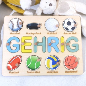May include: A wooden puzzle with a light brown background and colorful letters spelling the name "GEHRIG". The puzzle features images of different sports balls, including baseball, hockey puck, golf ball, soccer ball, football, tennis ball, volleyball, and basketball.