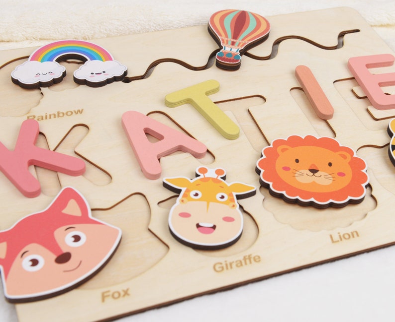May include: Wooden puzzle with colourful animal and rainbow pieces. The puzzle is shaped like the name "Katie" and includes the words "Rainbow", "Fox", "Giraffe", and "Lion".