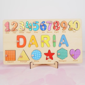 Op de afbeelding: Een houten puzzelbord met de naam DARIA in grote kleurrijke letters, de cijfers &eacute;&eacute;n tot en met tien met bijbehorende tekst, en zes geometrische vormen waaronder een vierkant, driehoek, cirkel, ster, zeshoek en hart met labels.