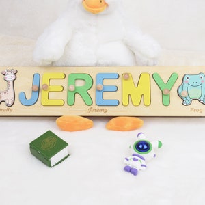 May include: A wooden name puzzle with the name "JEREMY" spelled out in colorful letters. The puzzle pieces are shaped like letters and have animal illustrations: a giraffe, a frog. The puzzle is on a white surface.