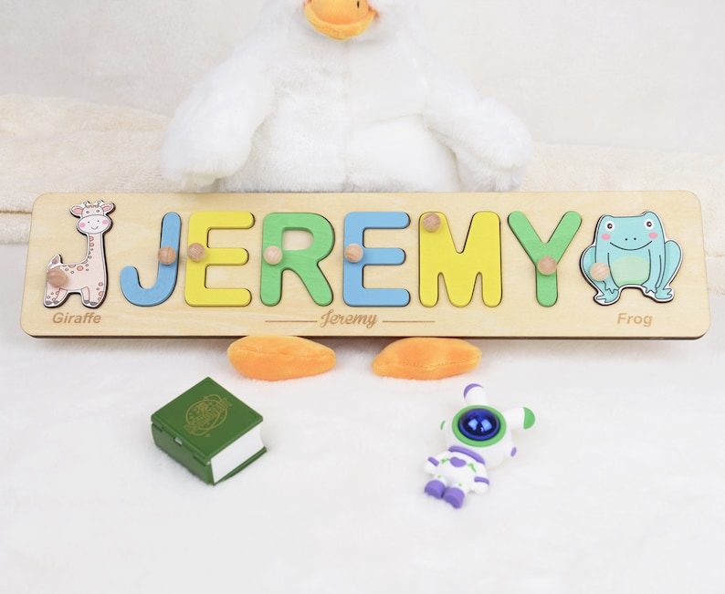 May include: A wooden name puzzle with the name "JEREMY" spelled out in colorful letters. The puzzle pieces are shaped like letters and have animal illustrations: a giraffe, a frog. The puzzle is on a white surface.
