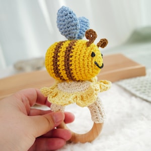 Op de afbeelding: Een gehaakt bij-rammelaar speelgoed met een houten ring. De bij is geel en bruin met een blauwe gehaakte vleugel. De rammelaar is een geweldig speelgoed voor baby's.
