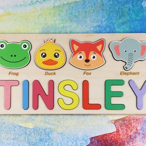 May include: A wooden personalized puzzle with the name "TINSLEY" spelled out in colorful letters. The puzzle also features four animal faces: a green frog, a yellow duck, an orange fox, and a gray elephant.